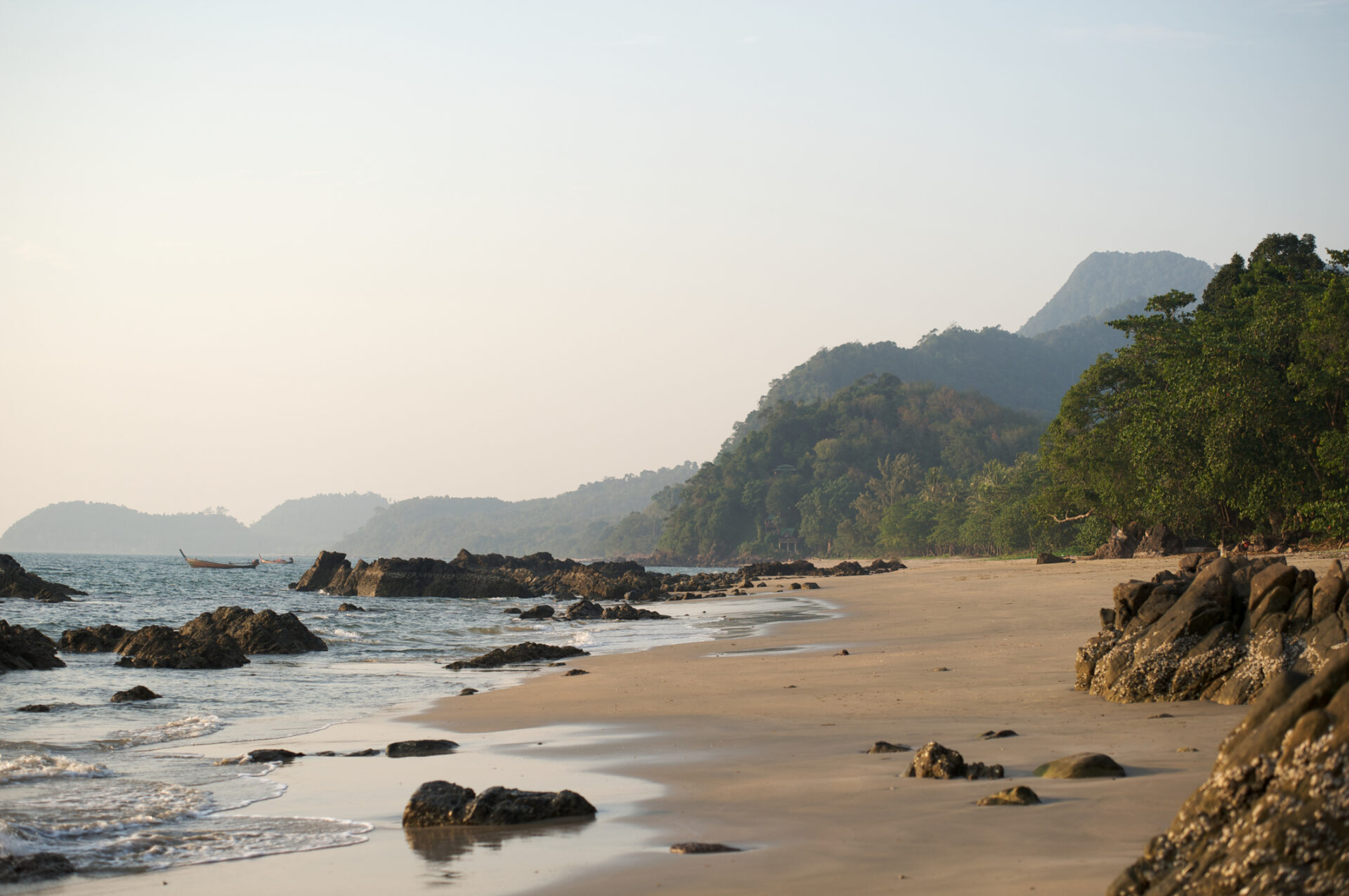 Klar havvand møder hvidt sand på en tropisk strand i Thailand, med palmer og blå himmel i baggrunden. Perfekt til ferie og afslapning.