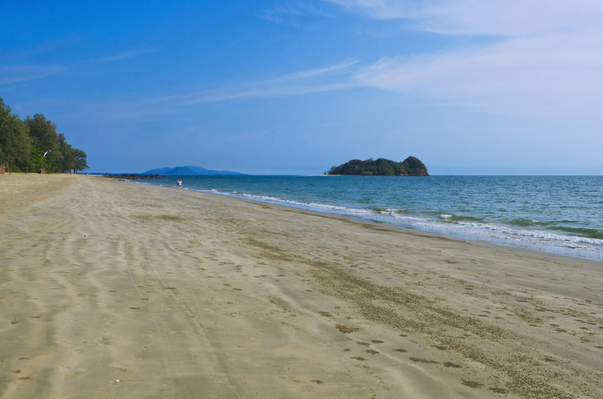 Idyllisk sandstrand på solbeskinnet dag i Thailand med krystalklart blåt vand, grønne palmer og lave bungalows omkranset af tropiske